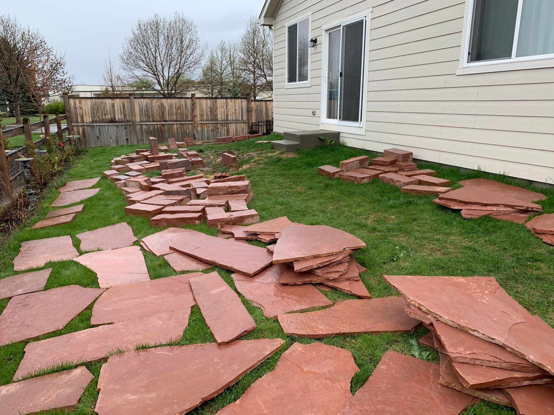 Red flagstone pavers scattered on green grass in a backyard, near a beige house and wooden fence.