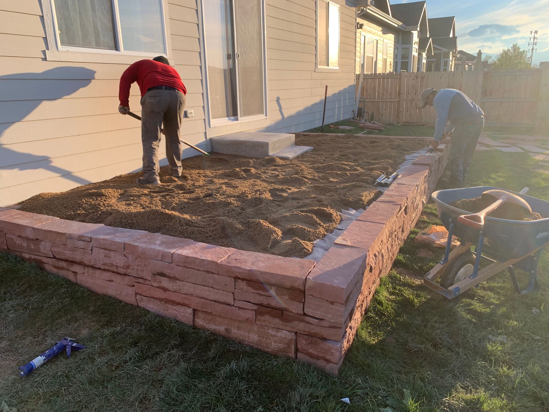 Two workers building a raised garden bed with red stone blocks. One shovels soil. Sunny backyard setting.