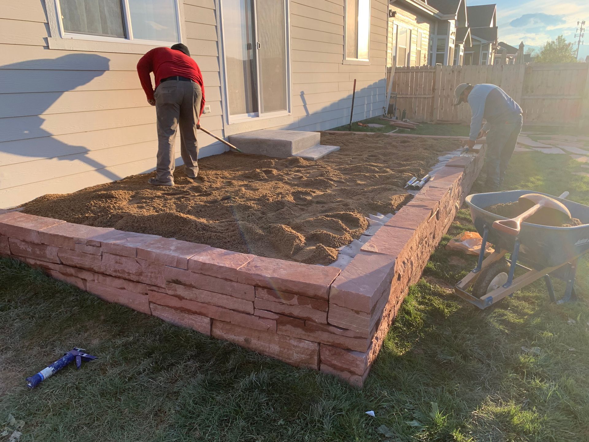 Men building a red brick planter in a backyard. One rakes dirt, another loads a wheelbarrow.