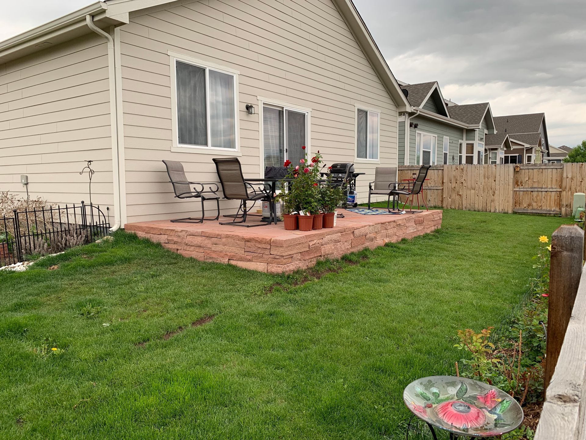 Backyard patio with chairs, grill, and rose bushes on a red brick platform, green grass, beige house, overcast sky.