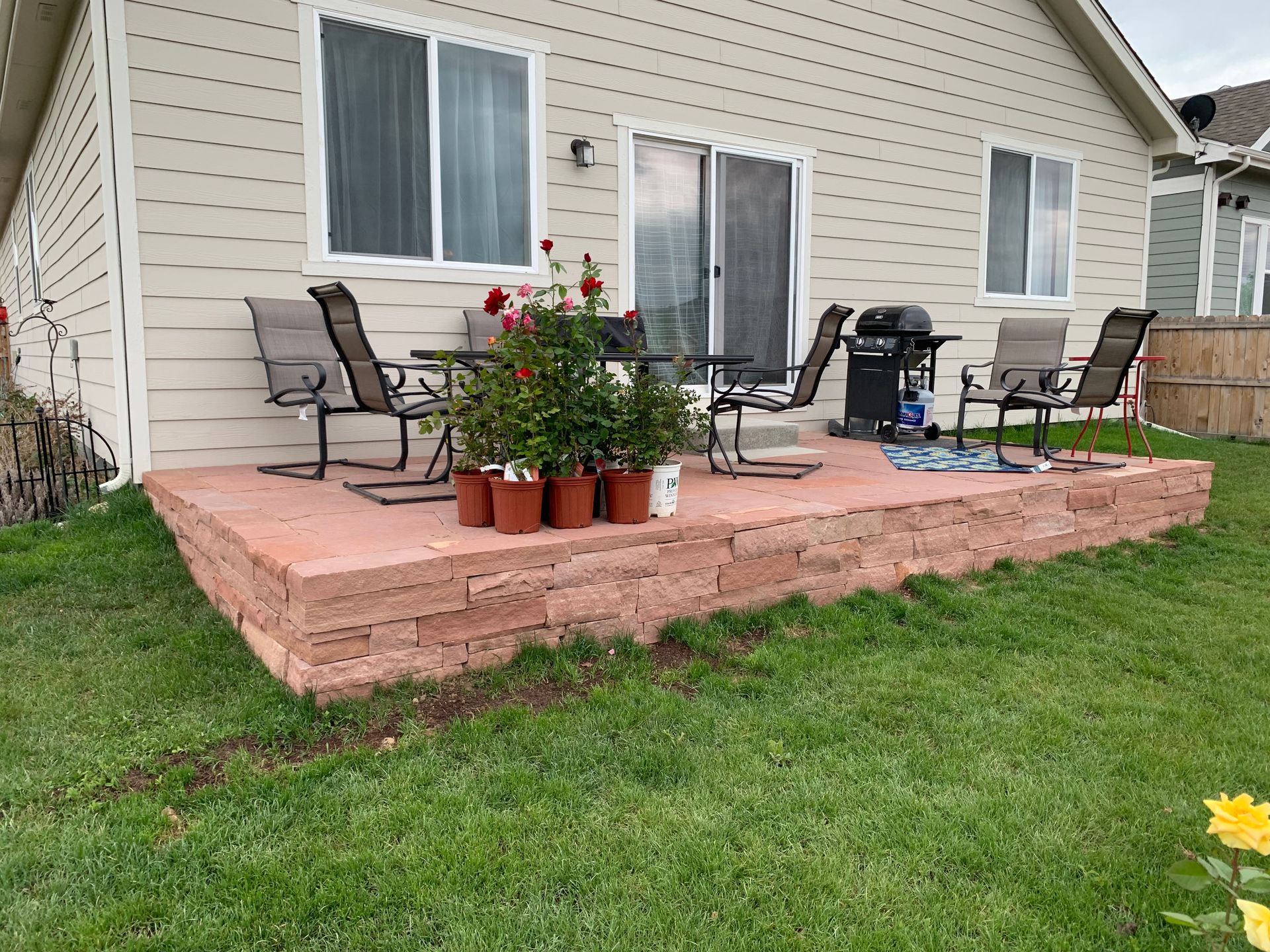 A backyard patio with red brick retaining wall, chairs, a grill, and potted plants.