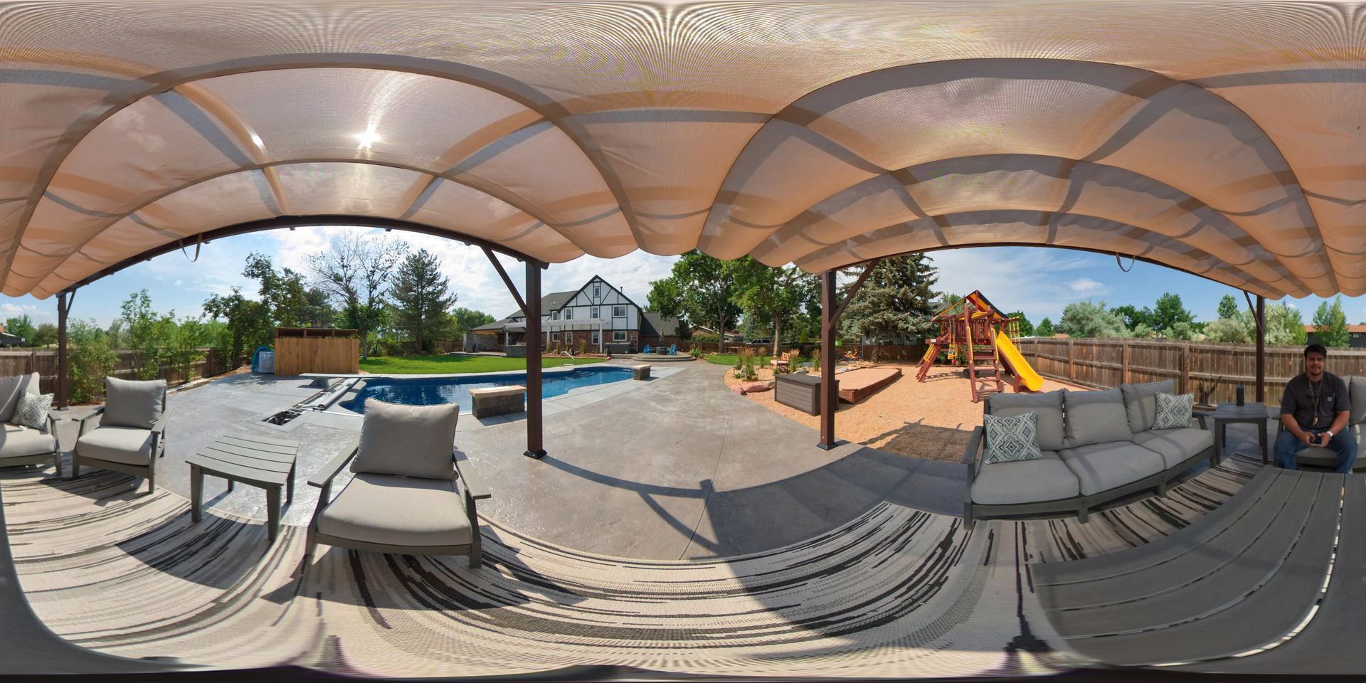 Backyard patio with pool, playground, and seating. Person sitting at the right. Overcast.
