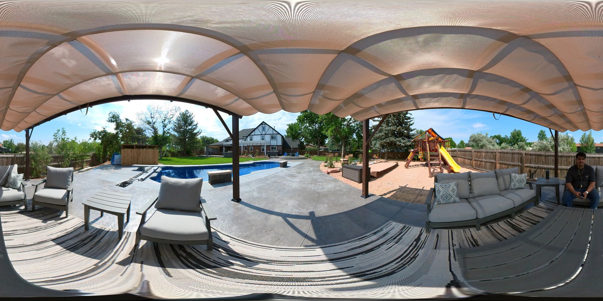 A backyard with a pool, seating area, and a playset under a canopy. A person sits on the right.