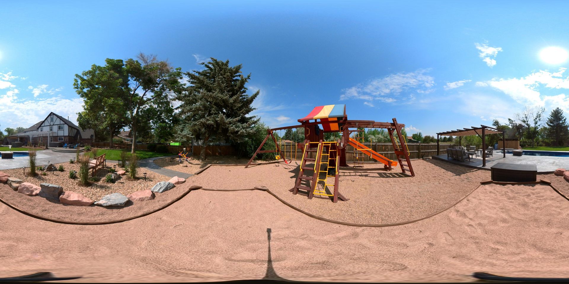 A playground with a slide and swings surrounded by sand, trees, and a house under a blue sky.