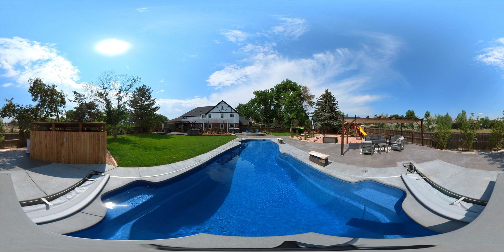 360-degree view of a backyard with a pool, a house in the background, a playground and blue sky.
