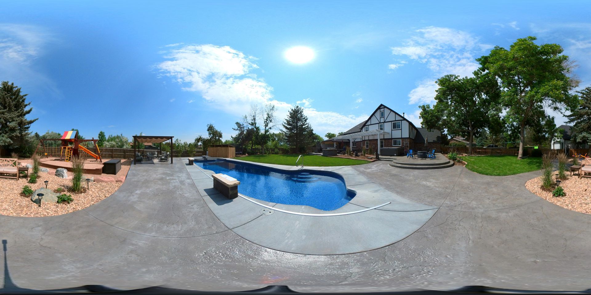 A sunny backyard with a pool, playground, and house. Blue sky with trees and gray concrete.