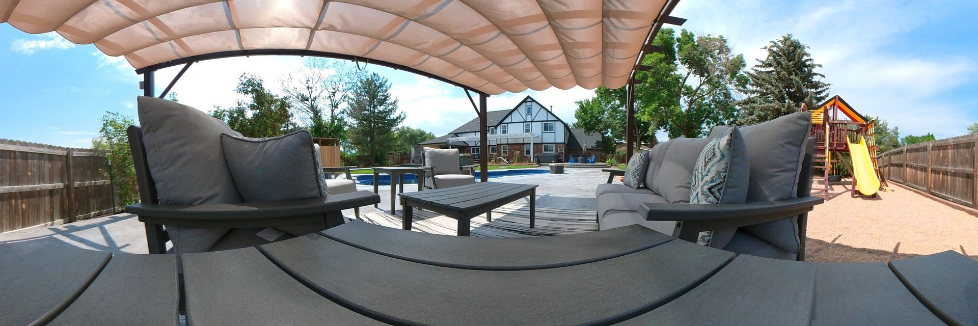 Outdoor patio with seating, pool, and playground, under a canopy on a sunny day.