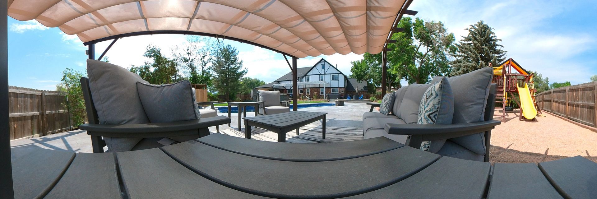 Patio with furniture under a canopy. Pool and playground in background, blue sky.