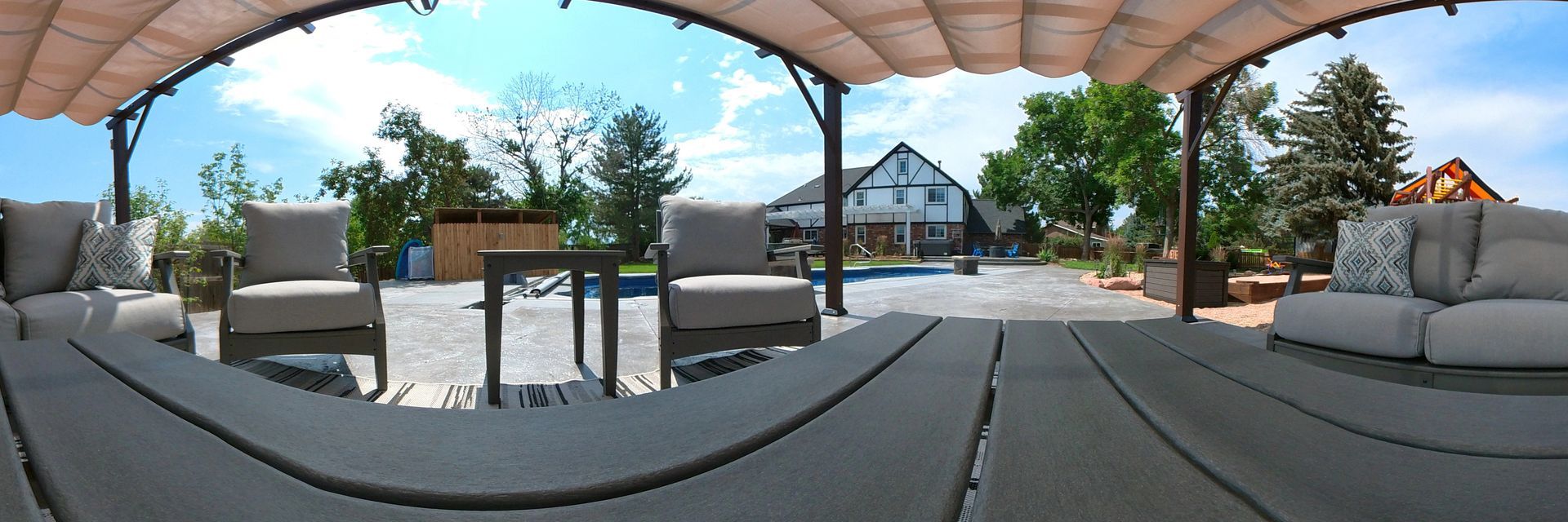 Outdoor patio with seating under a pergola, a house in the background. Sunny day.