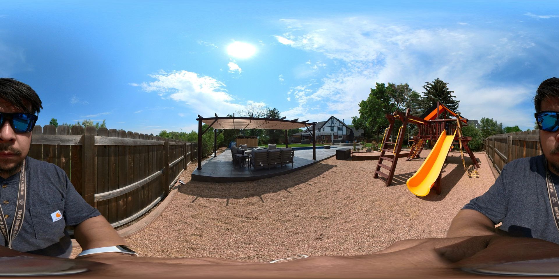 Man wearing sunglasses in a backyard with a playground, wooden fence, and patio under a blue sky.