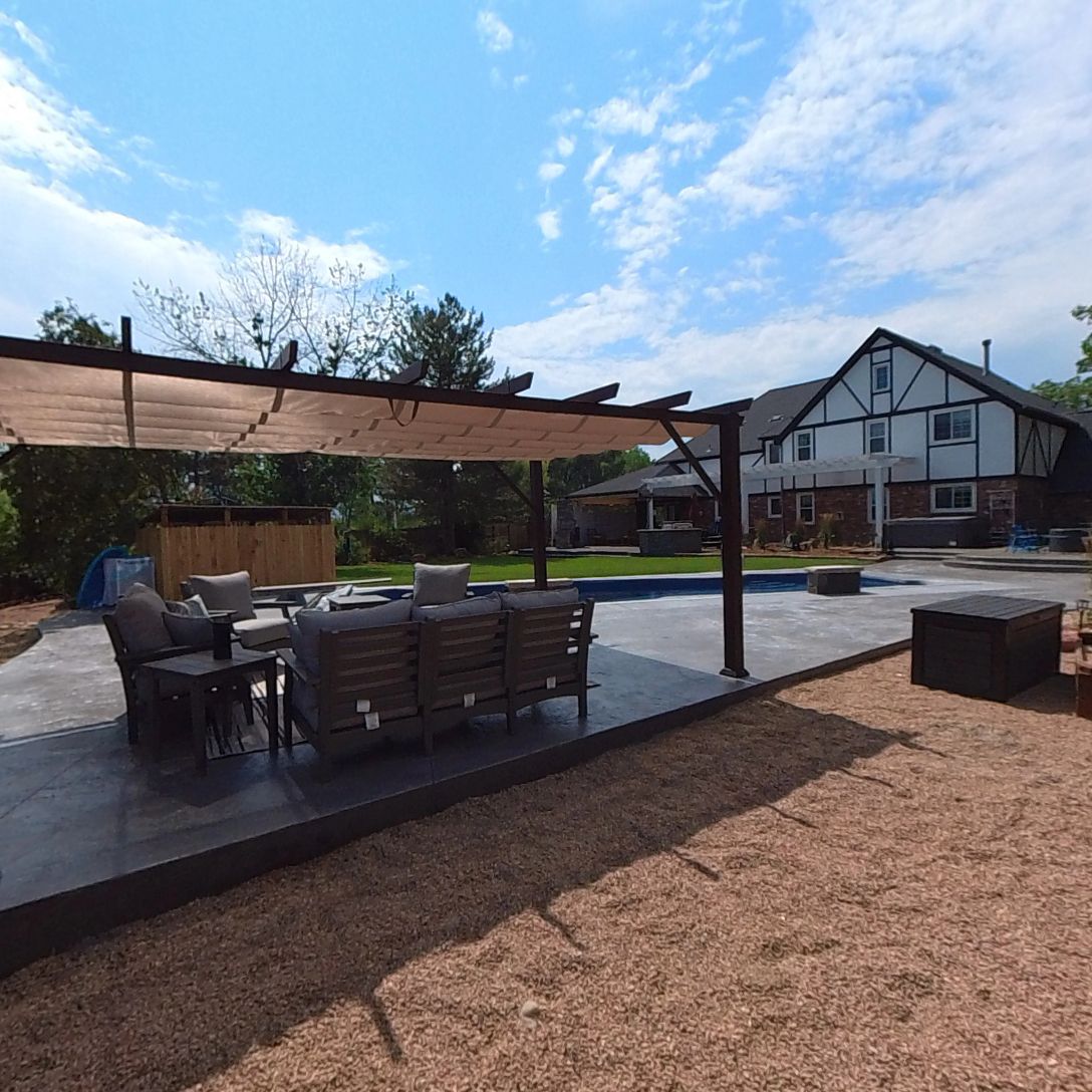 Outdoor patio with seating, pergola, pool, and house. Gravel ground, sunny day.