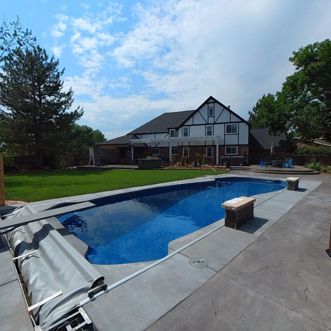 A backyard with a pool, concrete patio, and two-story house with black roof under a partly cloudy sky.