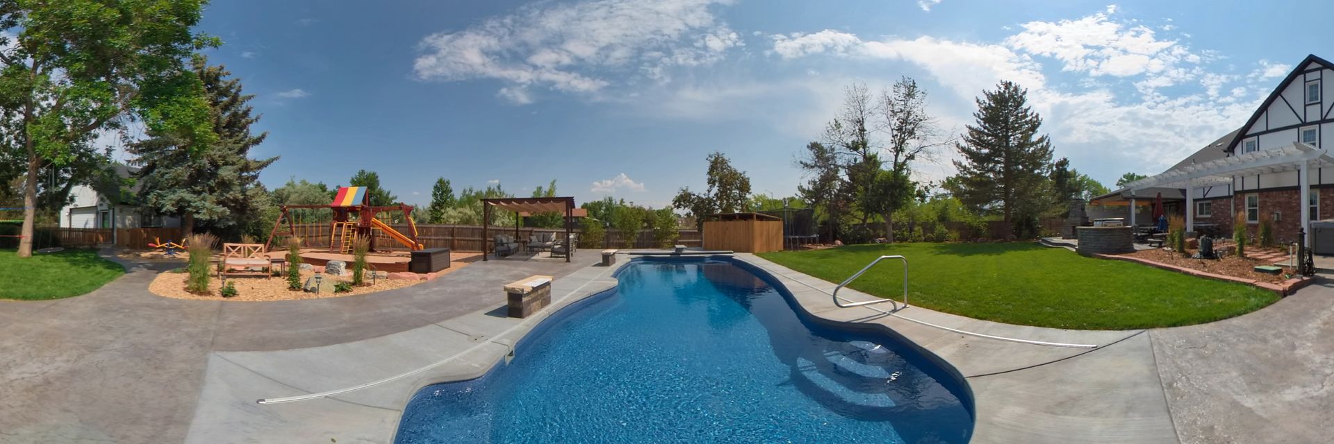 A backyard with a pool, lawn, and house under a blue sky.
