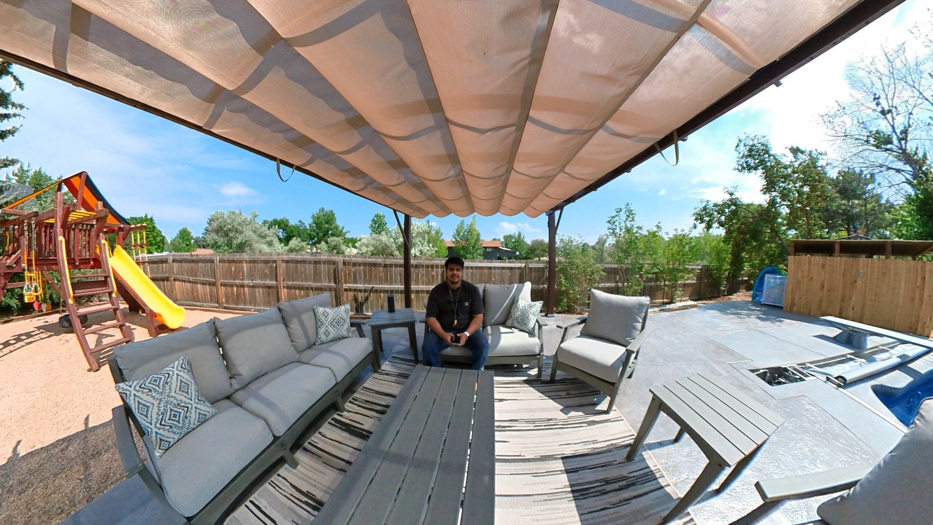 Man sitting on outdoor sofa beneath a canopy in backyard. Surrounded by furniture, play set, and fence.