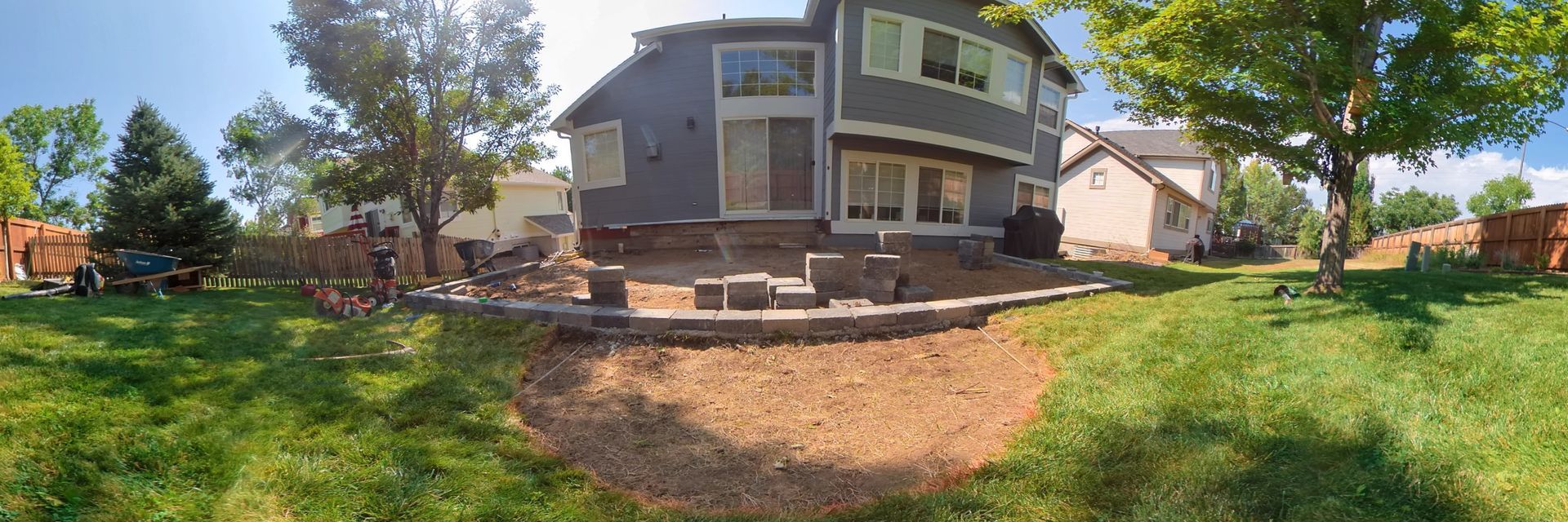 Backyard with a two-story gray house, patio, and lawn. Brown mulch bed and trees in the sunny scene.