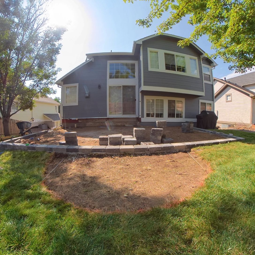 Backyard view of a two-story gray house. A patio is under construction, and the lawn is partially seeded. Sunny day.