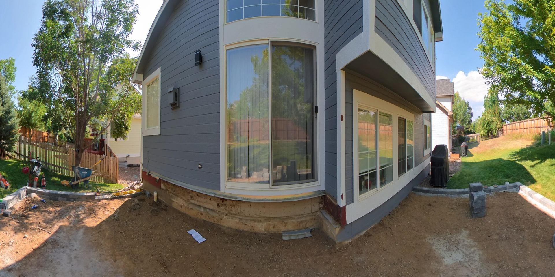 Exterior view of a two-story gray house with windows and a large sliding glass door, surrounded by dirt and grass.