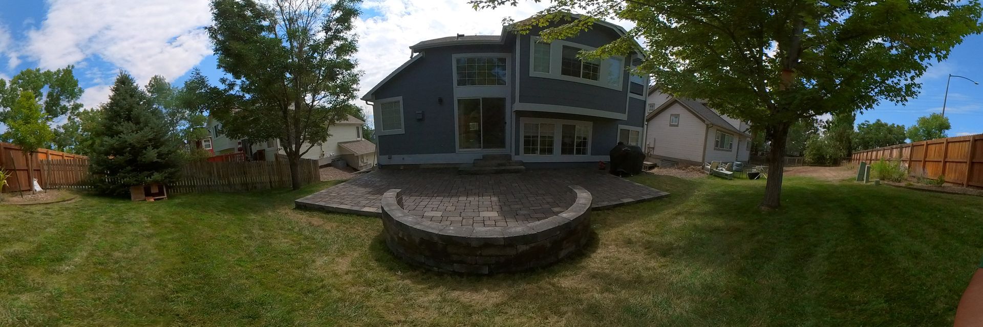 Backyard with grass, patio, and a two-story blue house. A fire pit is in the center. Bright sunny day.