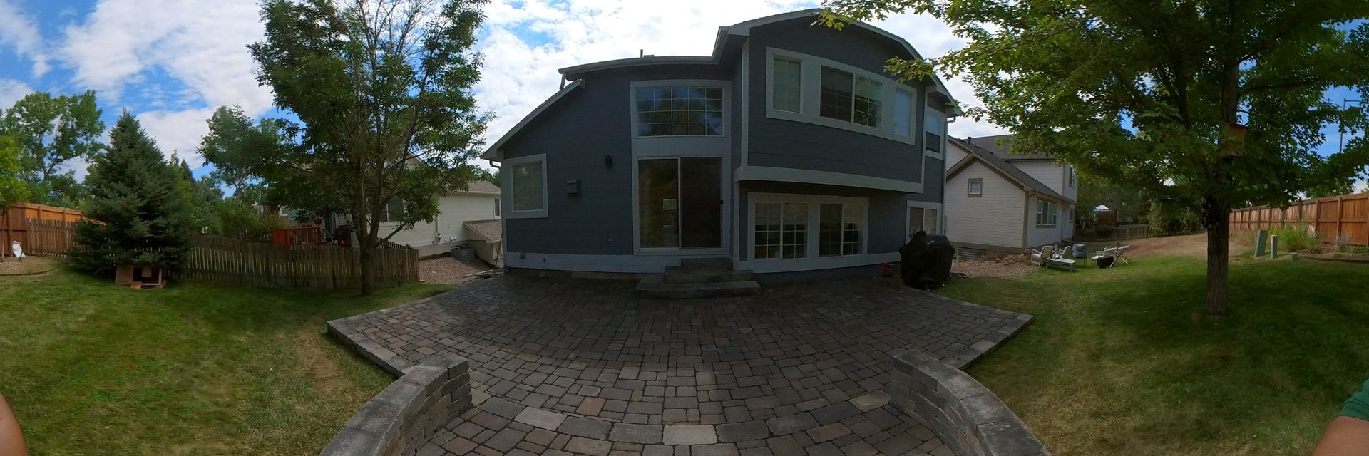 Backyard of a two-story blue house with a stone patio, green grass, and trees. Blue sky with clouds.