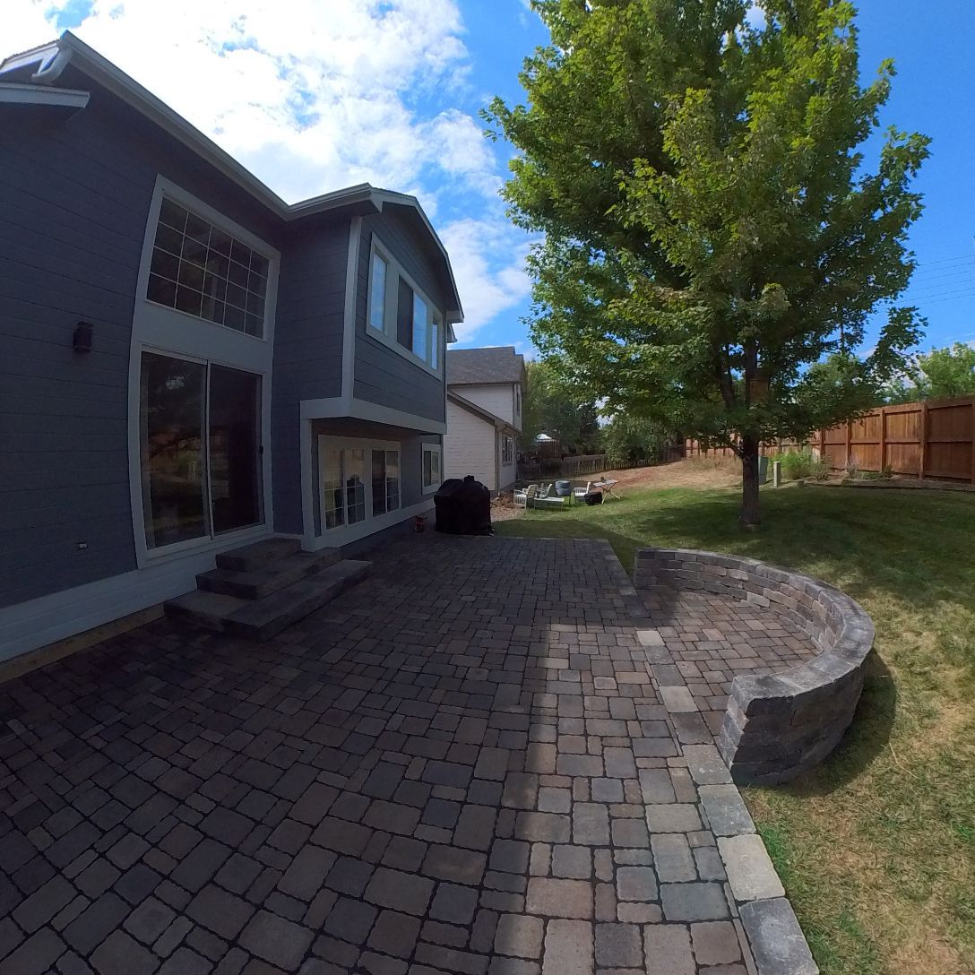 Backyard patio with gray brick, house, tree, and blue sky.