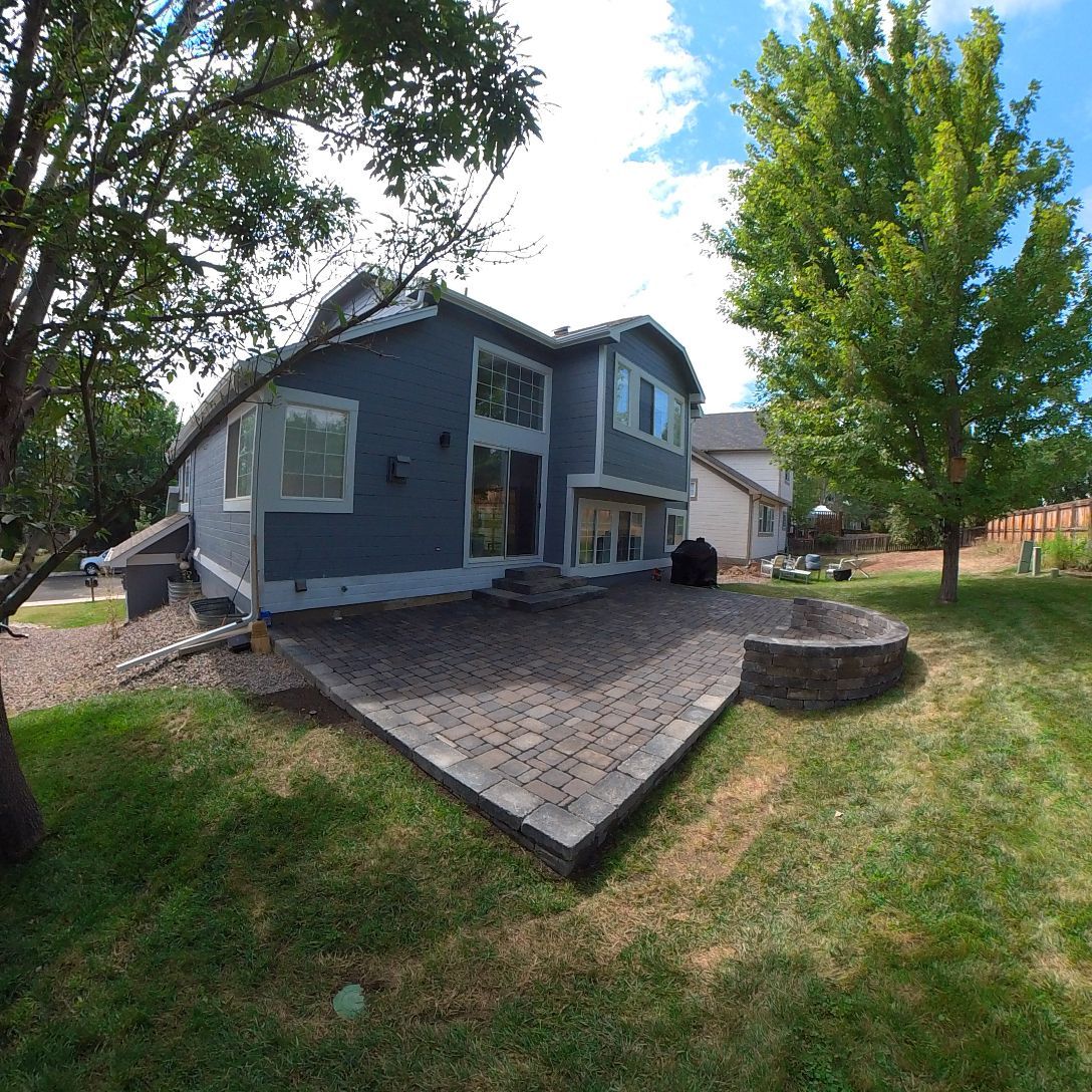 Backyard with a blue house, patio, green grass, and trees under a cloudy sky.