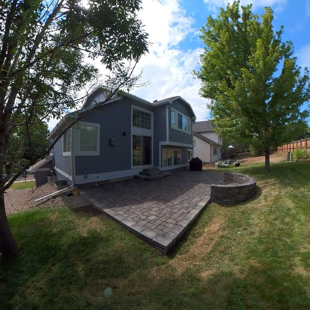 Backyard with gray house, patio, and green lawn under a blue sky.