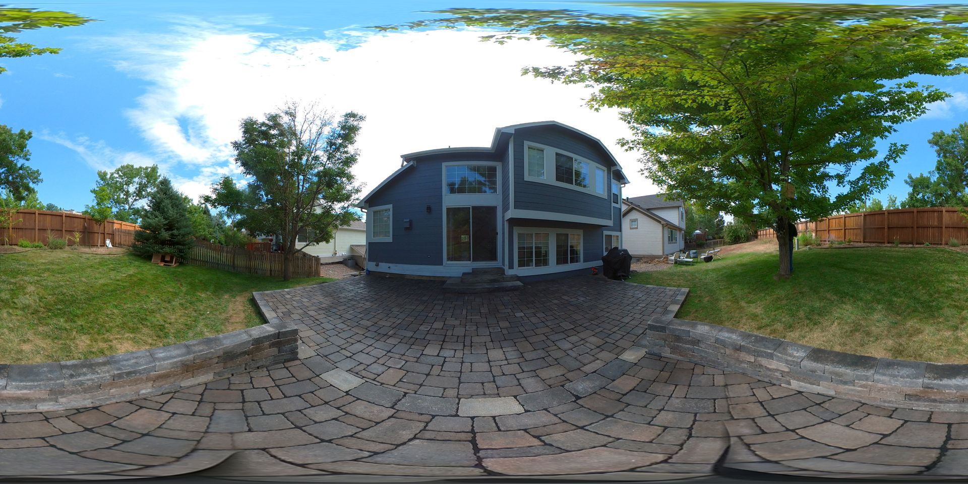 A two-story blue house with a curved roof. Large tree to the right, blue sky, and a brick patio.