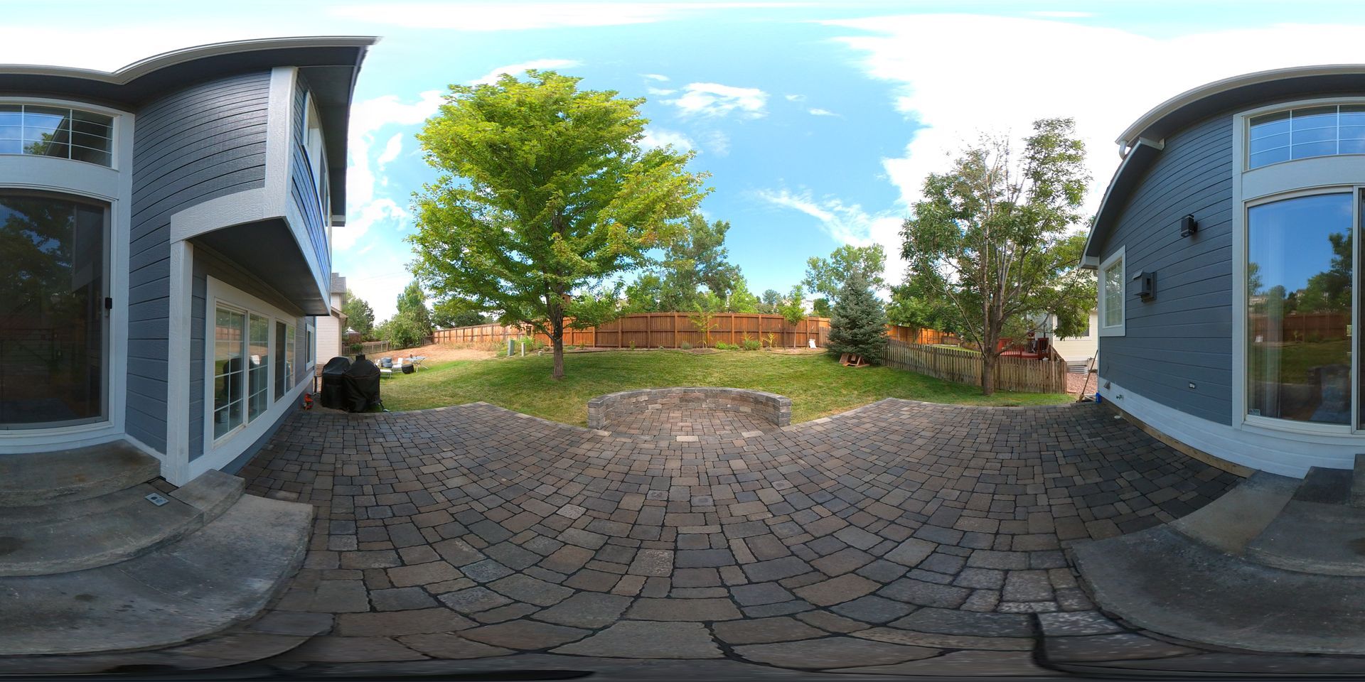 Backyard with a brick patio, trees, and a house with blue siding under a blue sky.