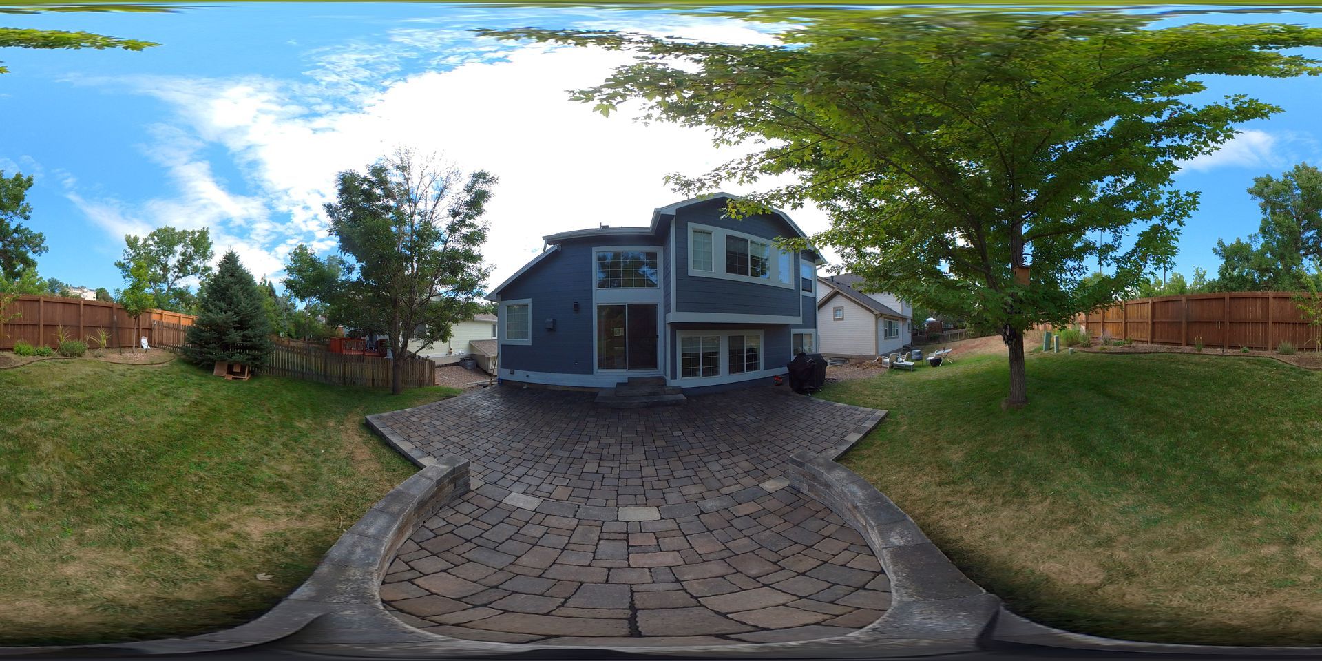Backyard view of a two-story blue house with a brick driveway and green lawn under a blue sky.