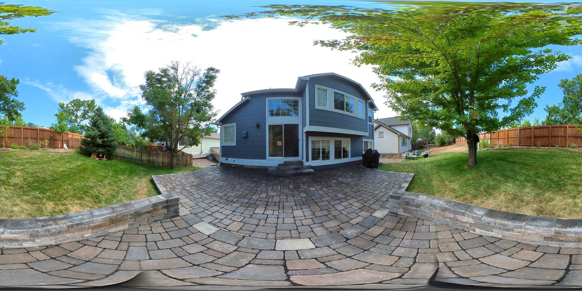 A blue house with a curved roof sits on a brick patio, surrounded by green grass and trees under a blue sky.