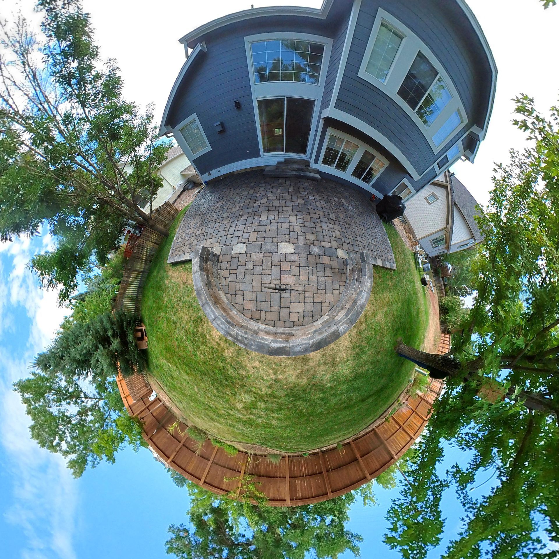 Tiny planet view of a backyard with a blue house, brick patio, green grass, and a wooden fence.
