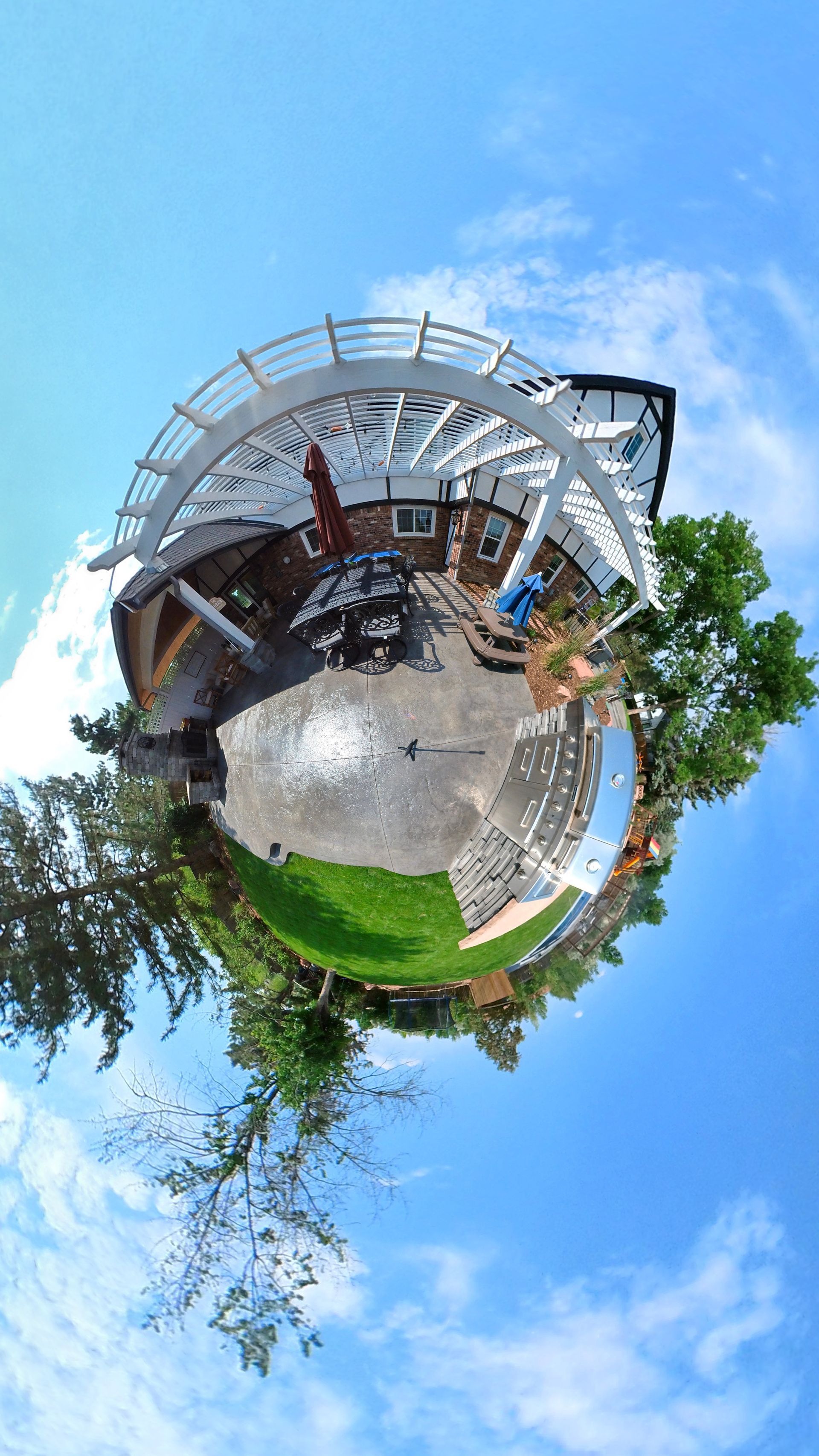 Tiny planet view of a backyard with a pergola, grass, patio furniture, and blue sky.