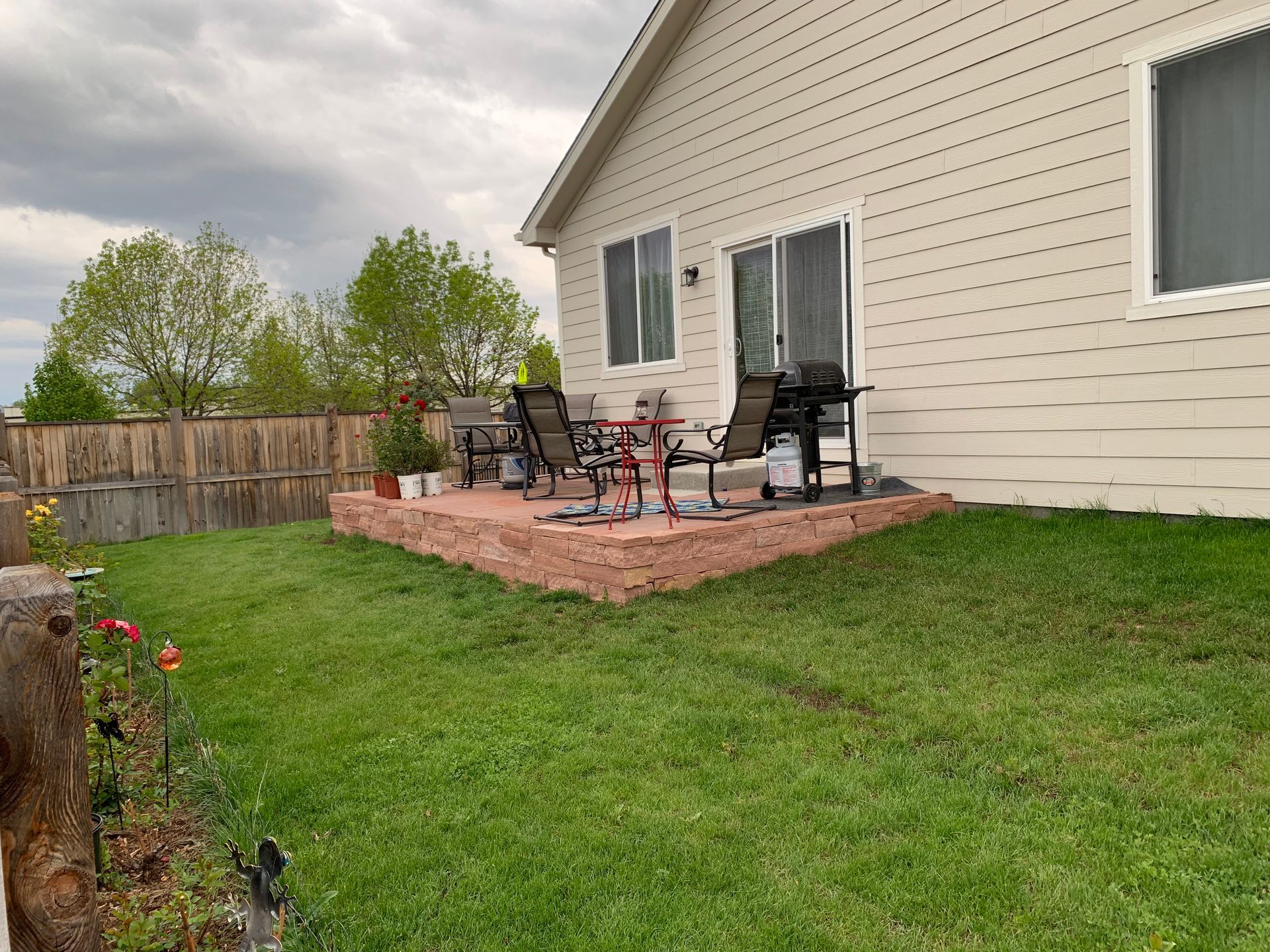 A backyard patio with a grill, chairs, and table. Green grass, a wooden fence, and a beige house are visible.