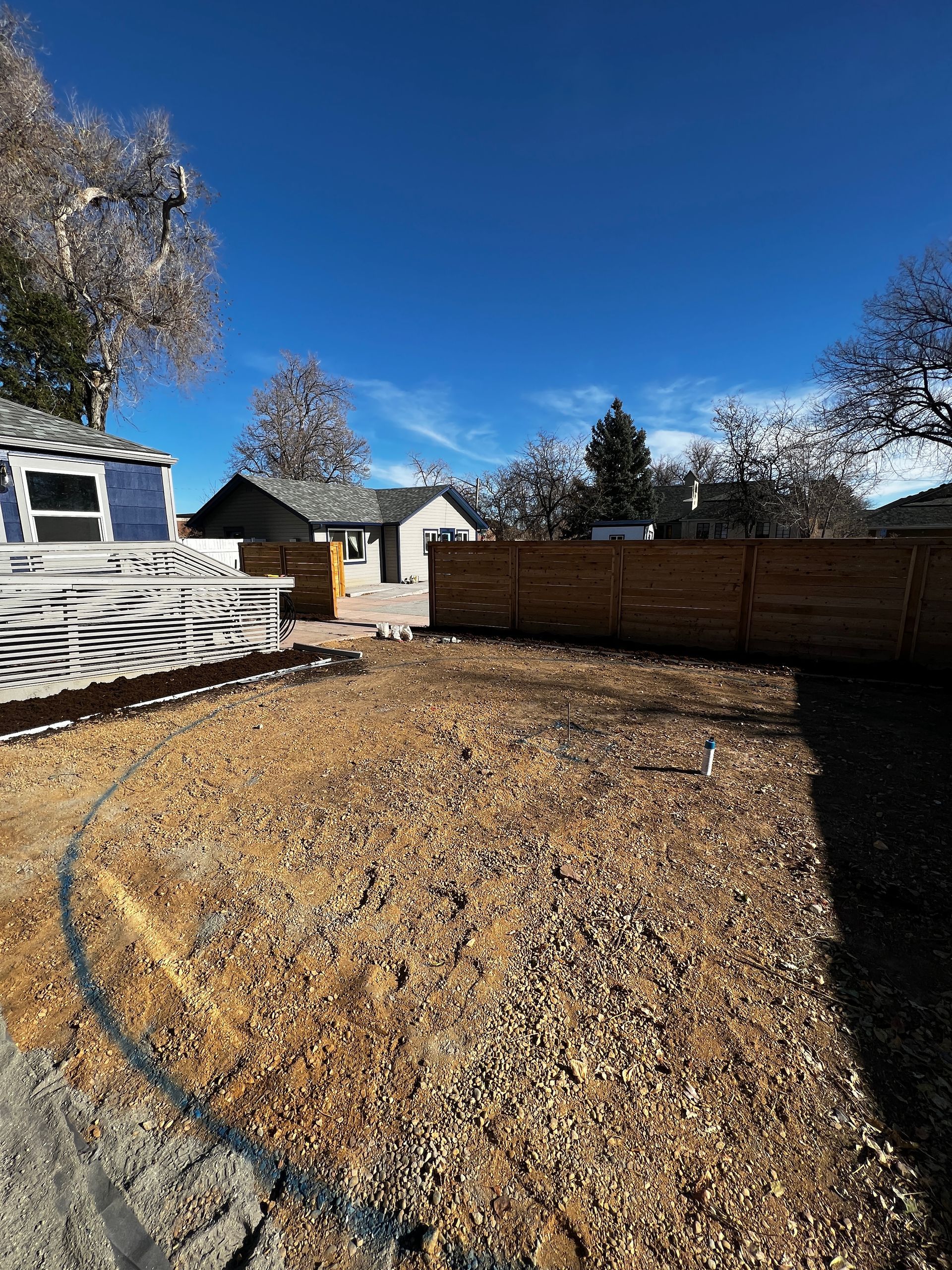A vacant lot covered in gravel, with a fence and houses under a blue sky.
