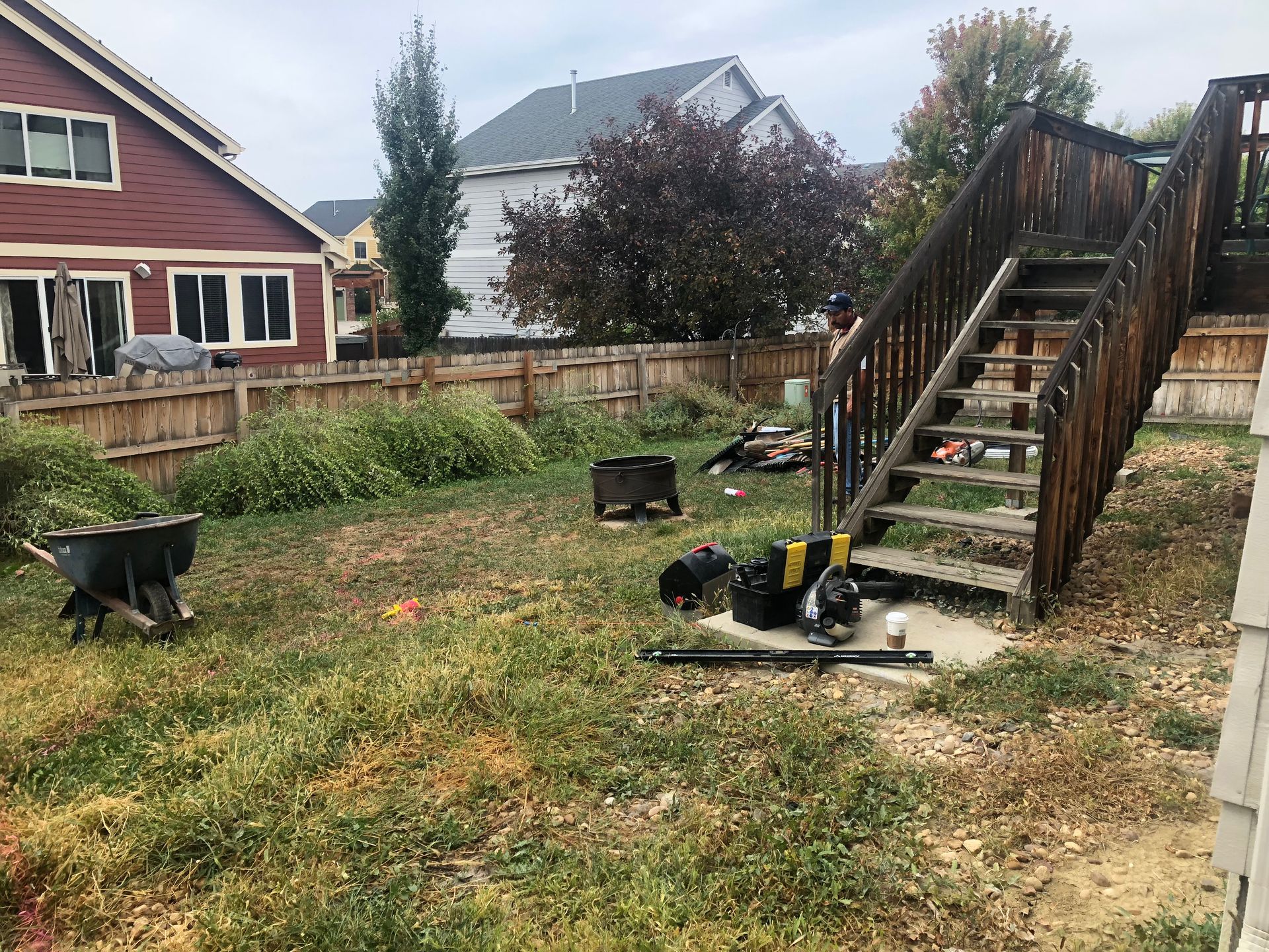 Backyard with stairs, fence, overgrown grass, wheelbarrow, and assorted debris.