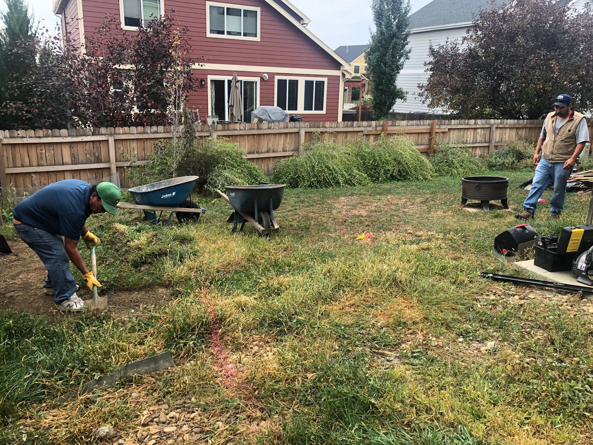 Two men working in a weedy backyard with tools, wheelbarrows, and a red house in the background.