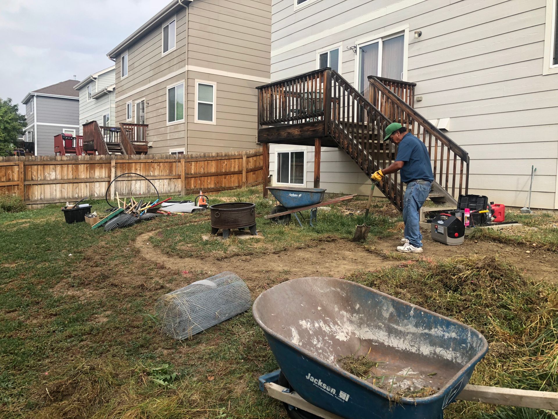 Man working in backyard near a deck and fence, using tools and wheelbarrows to clear debris.