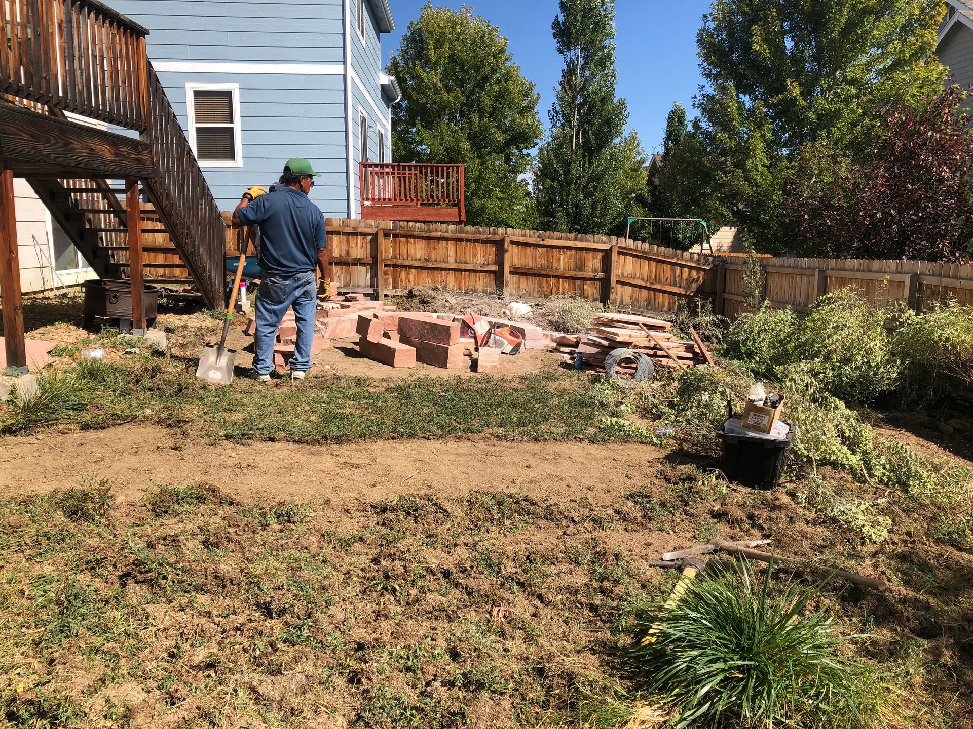 Man working in a backyard, clearing debris and bricks near a wooden fence and deck.