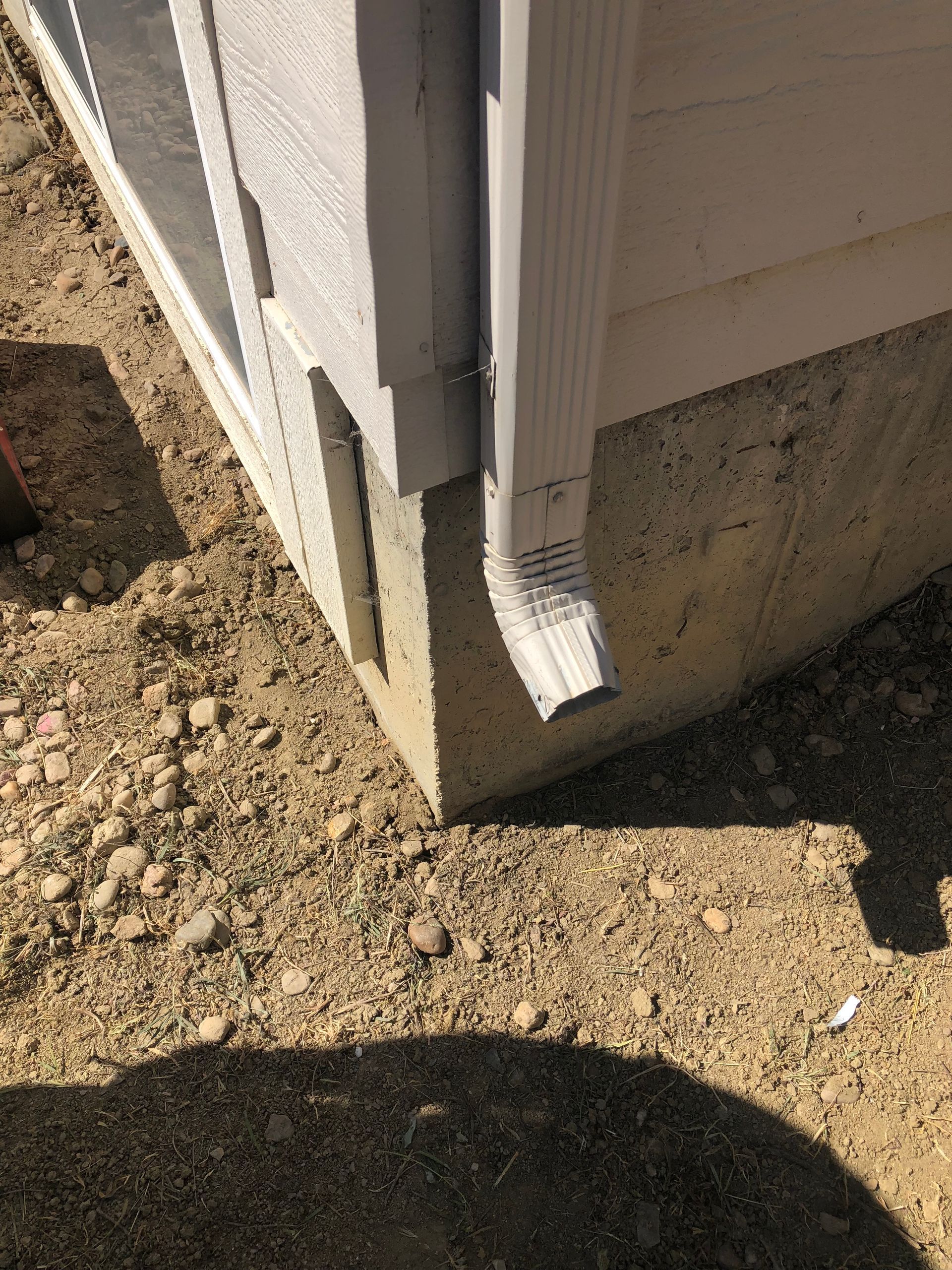 Gutter and downspout on a light-colored building with tan dirt and gravel at its base.