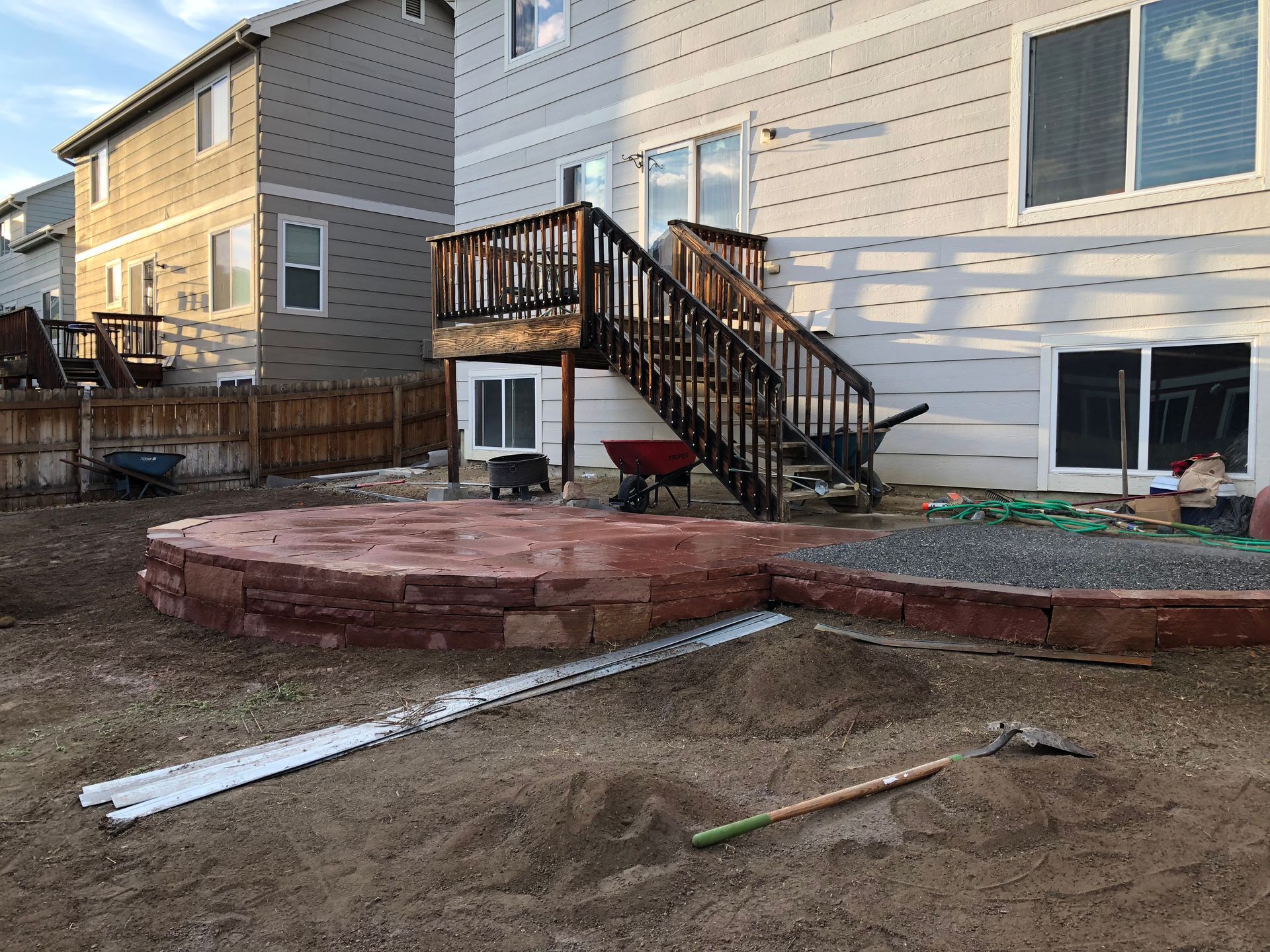Backyard with a raised red brick patio and wooden deck. Gravel, dirt, and construction tools are visible.
