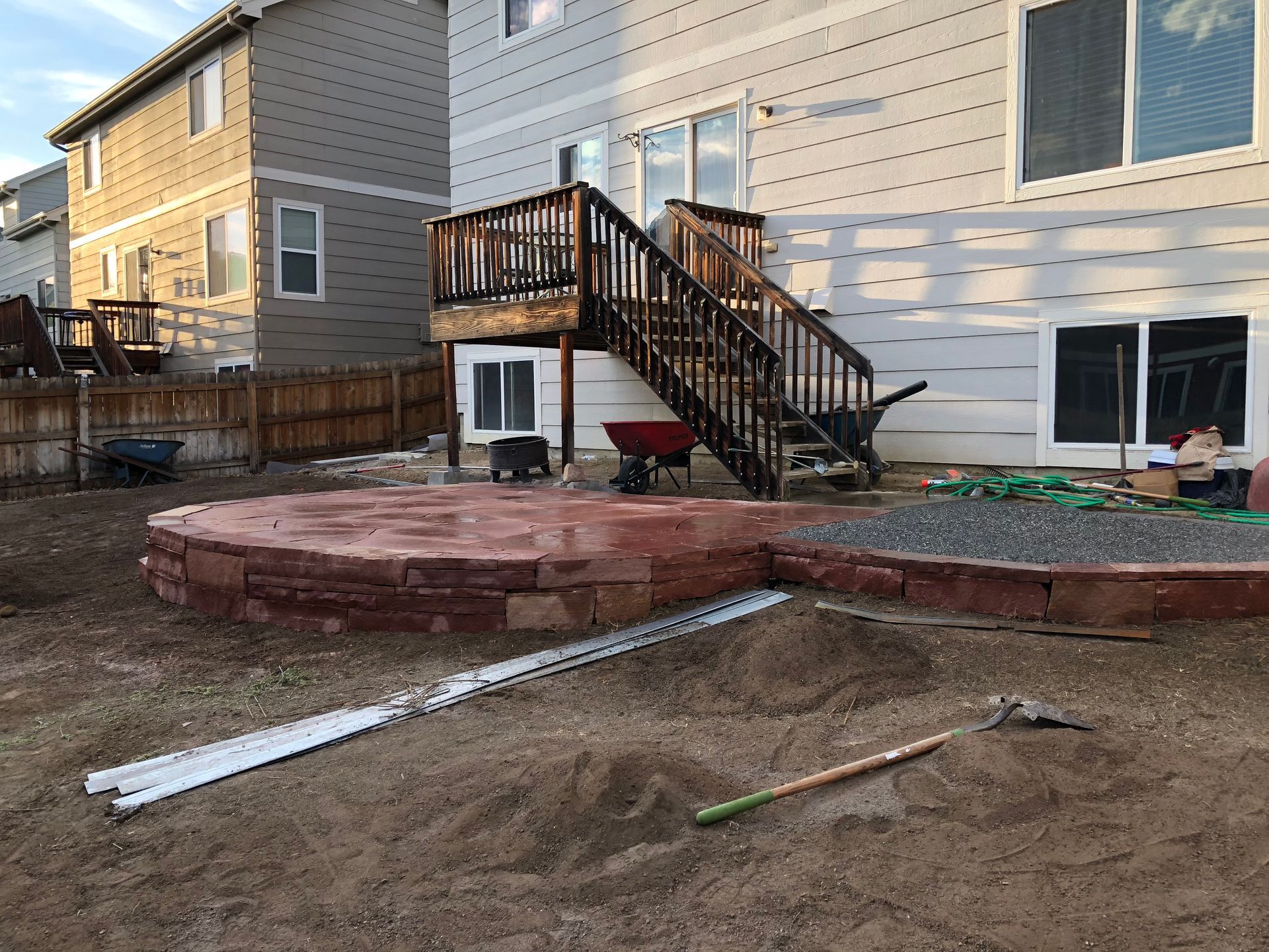 Backyard with a raised, reddish-brown patio, wooden deck with stairs, and a house in the background.