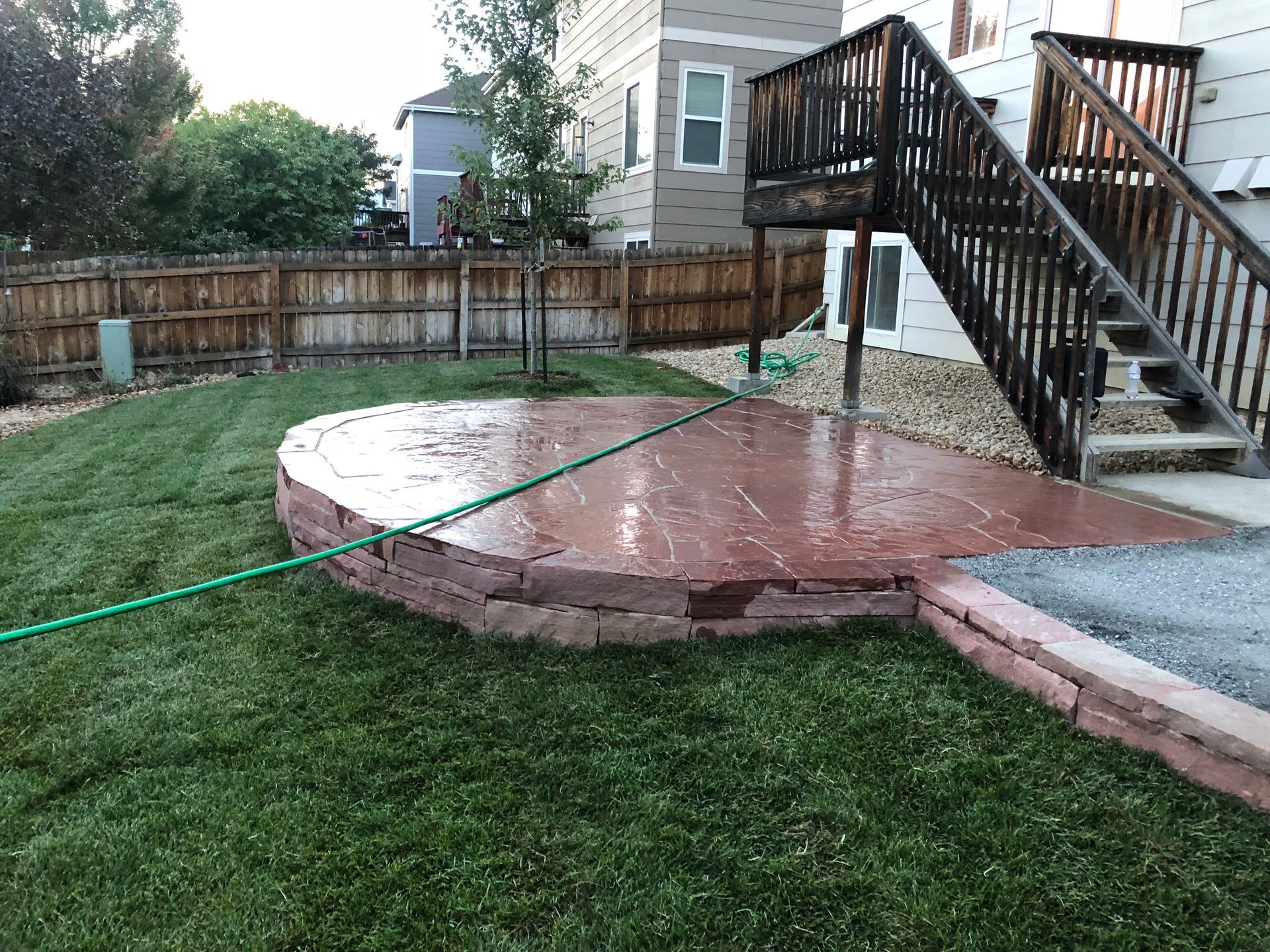Backyard with a red stone patio, green grass, and wooden stairs.