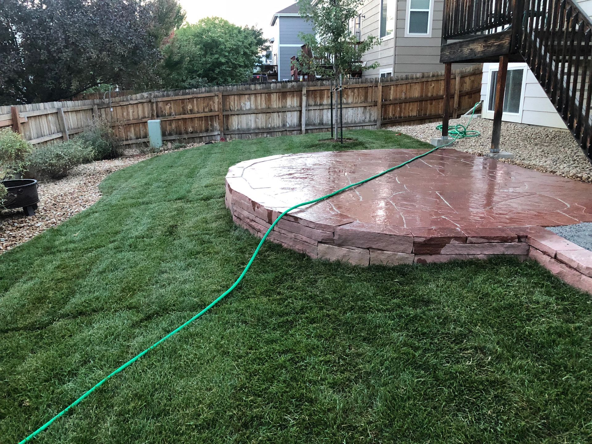 Green hose on a red stone patio, wet green grass in a backyard with wooden fence.