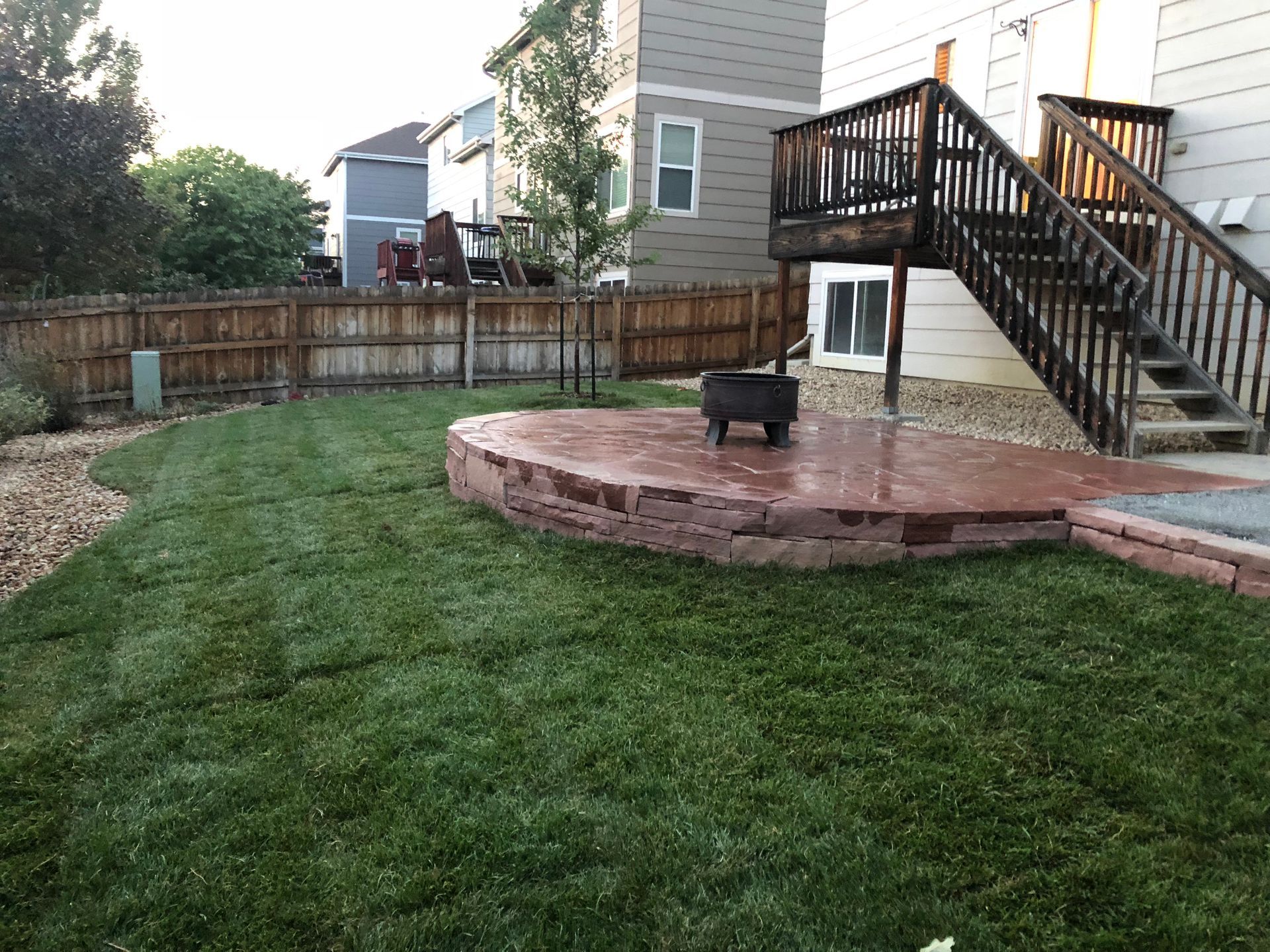 Backyard with a brick patio, fire pit, green grass, wooden fence, and stairs leading to a deck.