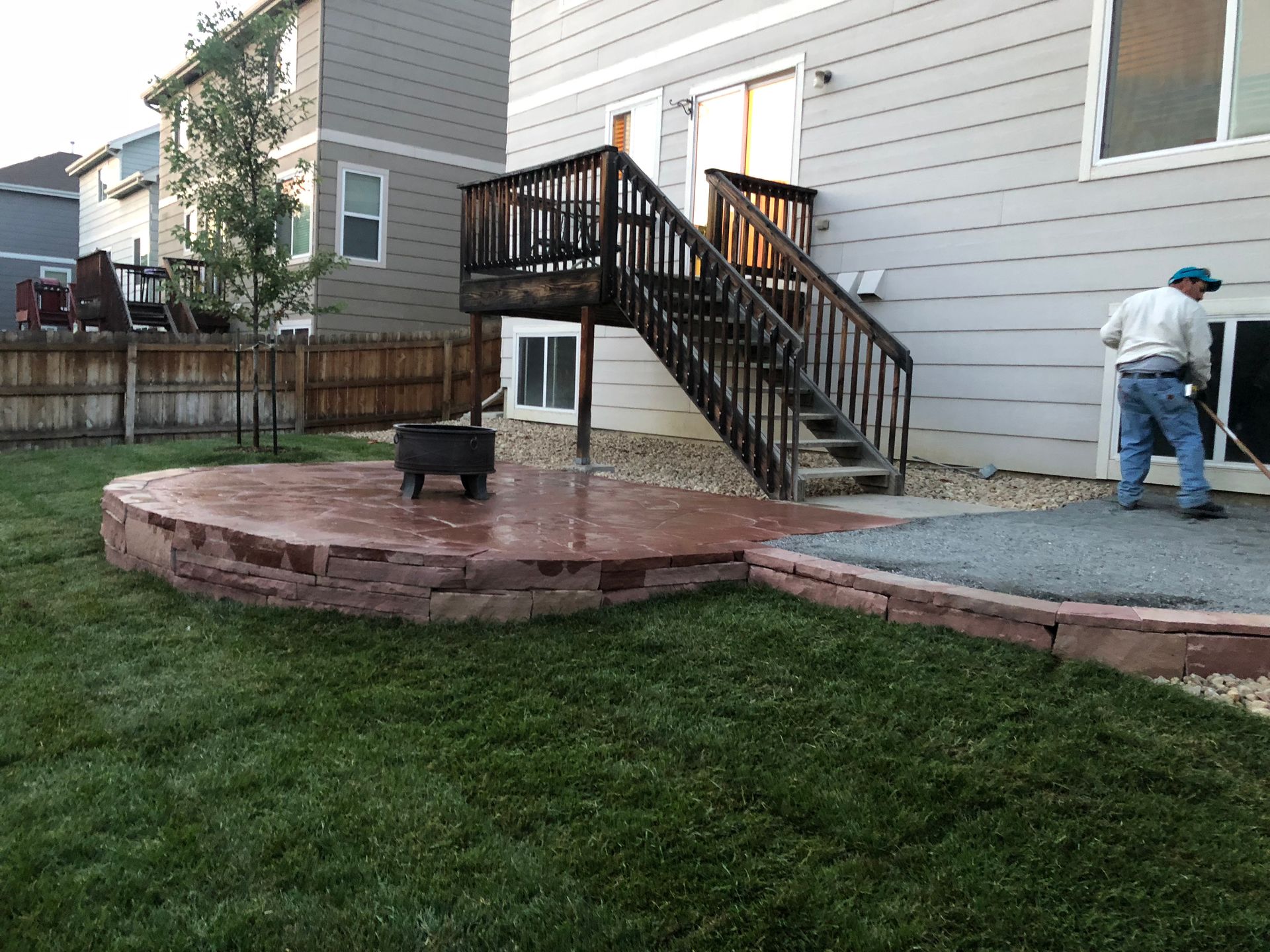 Backyard with a raised red concrete patio, wooden deck, person sweeping.
