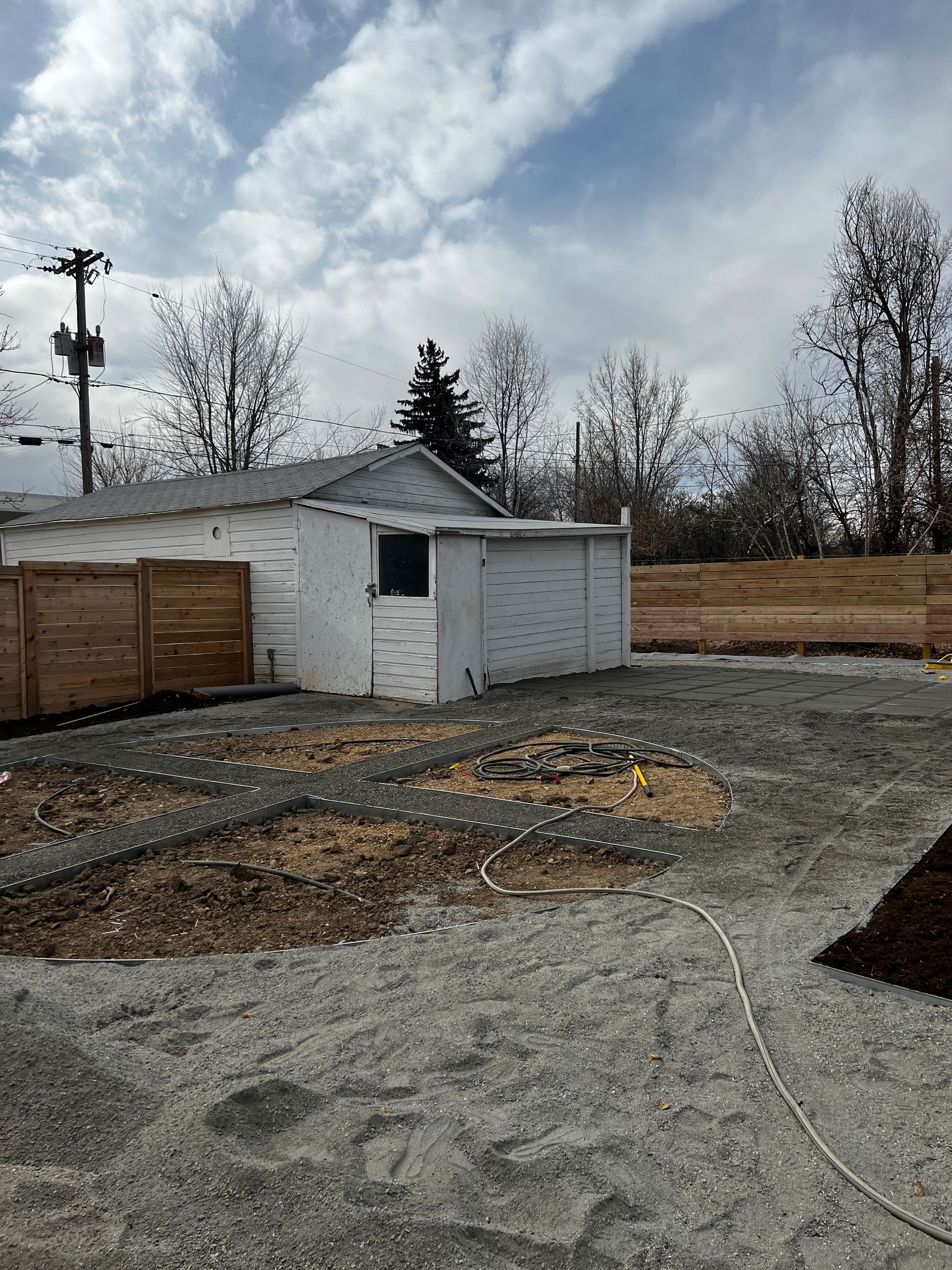 Gravel yard with a white shed, brown fence, and trees under a cloudy sky.