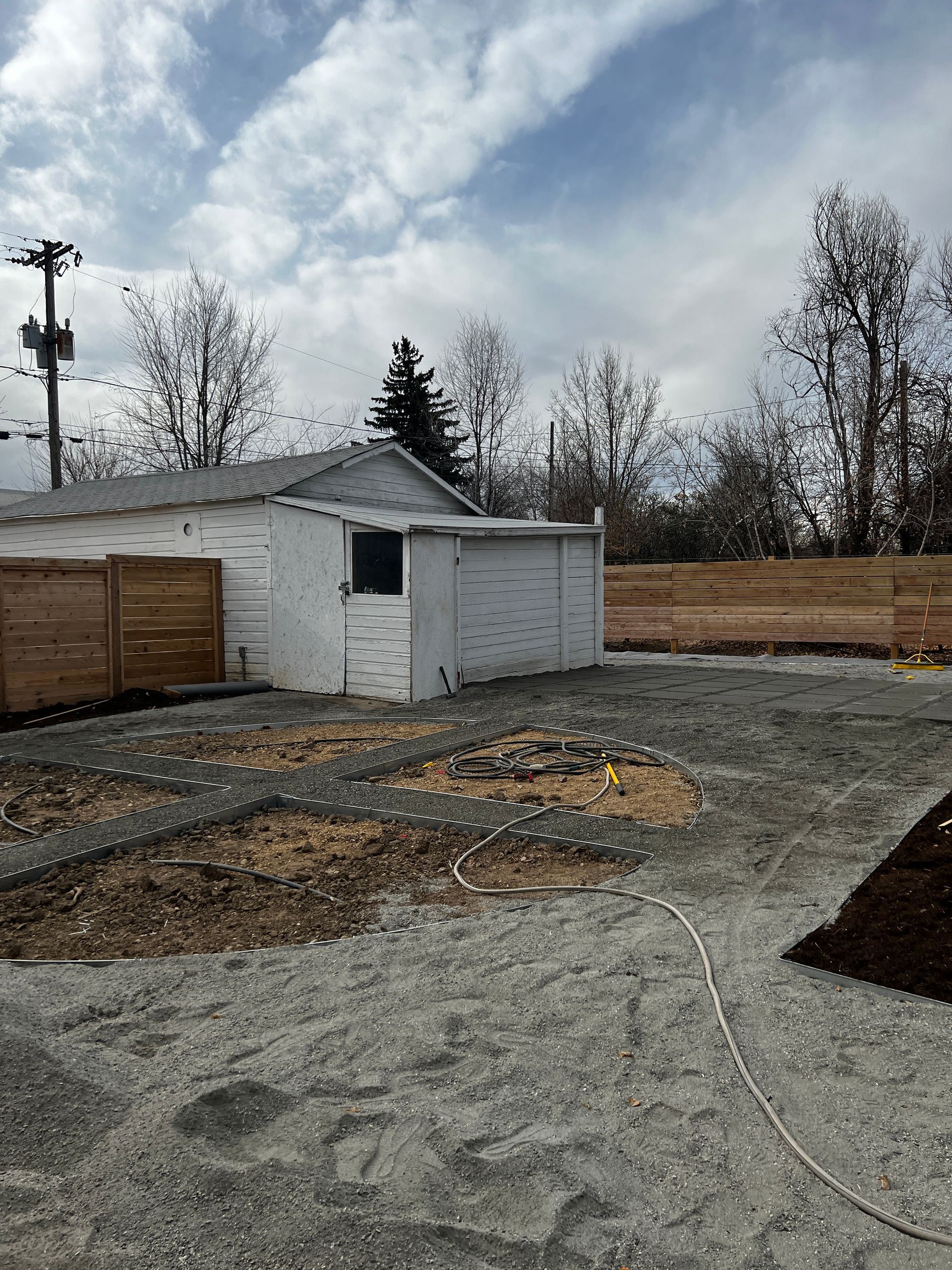 A gravel yard with a shed and wooden fence. Cloudy sky.