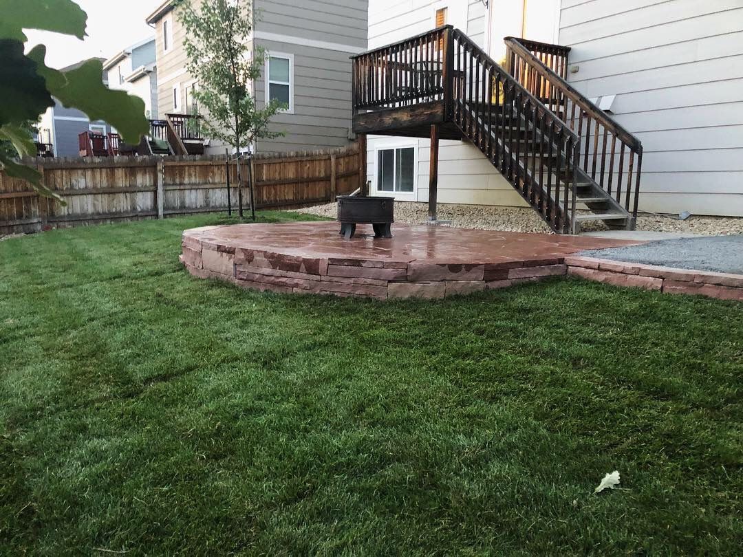 Backyard with circular brick patio, fire pit, stairs to a deck, and green lawn.