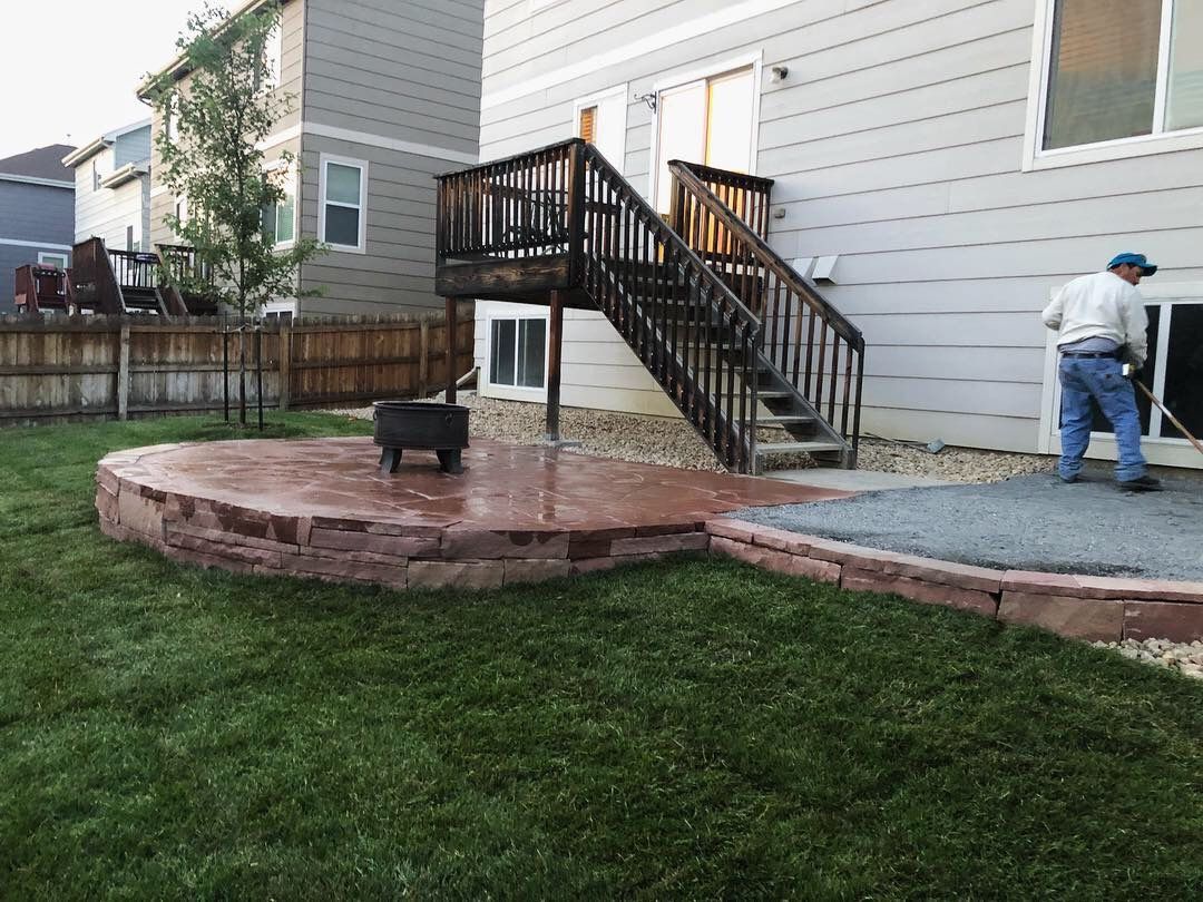 Backyard with a brick-rimmed patio, stairs to a deck, and a person working near a sliding glass door.