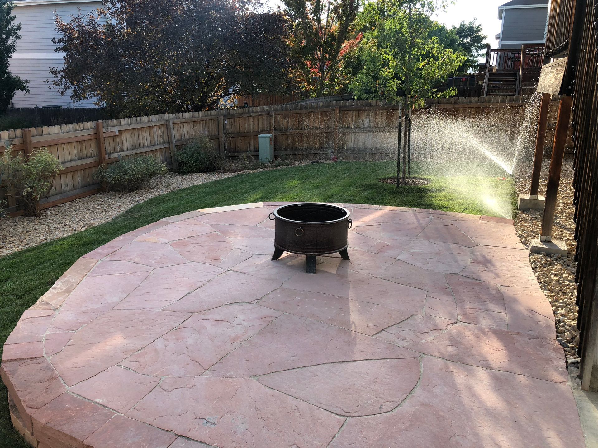 Backyard patio with fire pit, sprinkler spraying water, green grass, and wooden fence.